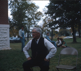 Actor wearing black suit vest seated outside of courthouse building. Additional actors seated in background.